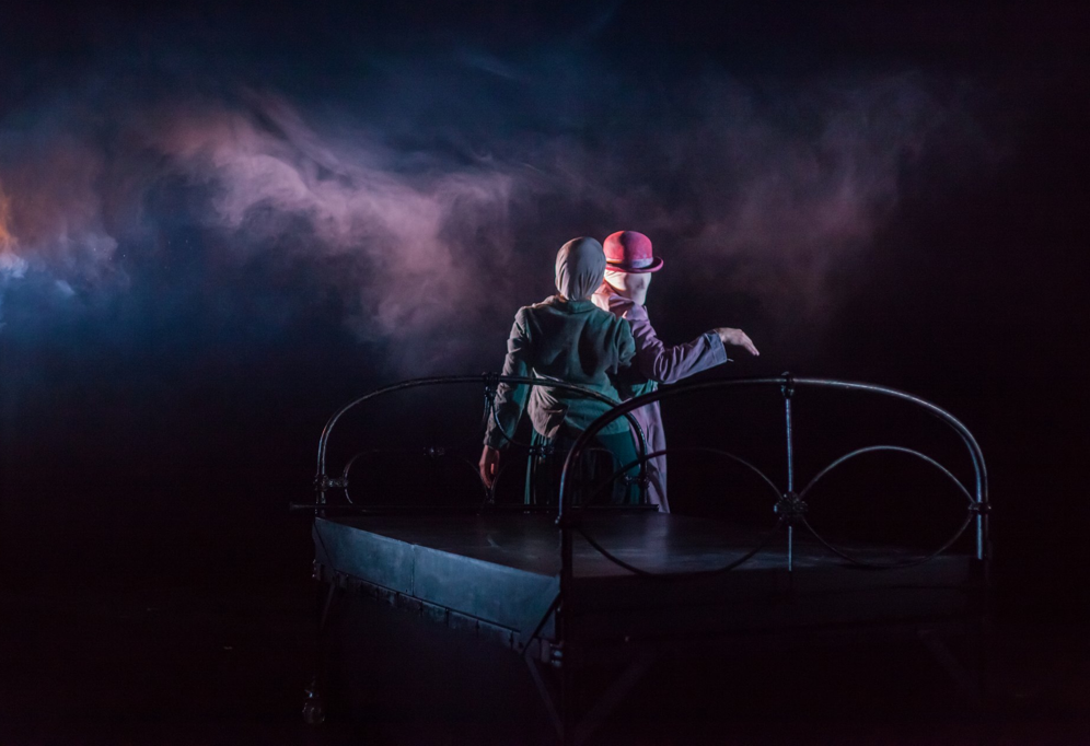 Image: A dark stage, with swirling fog highlighted in reds and purples. Onstage is a large, iron wrought empty bed. Behind the bed there s a woman with her back to the audience and in front of her is a man with cloth on his face looking over his shoulder to the audience.